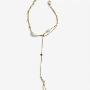 May include: A gold-toned chain hand bracelet featuring two light blue spherical beads and two small star-shaped charms. The jewellery piece includes a delicate chain that loops around a finger, connecting to a wrist chain with a lobster clasp closure.