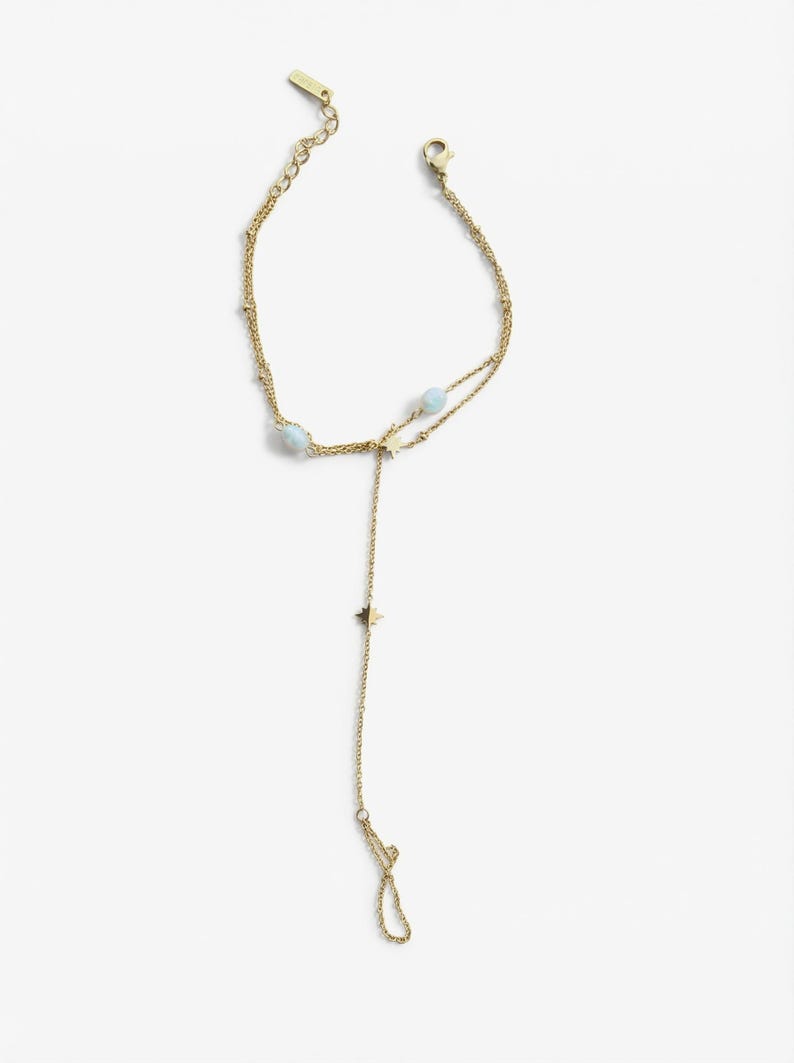 May include: A gold-toned chain hand bracelet featuring two light blue spherical beads and two small star-shaped charms. The jewellery piece includes a delicate chain that loops around a finger, connecting to a wrist chain with a lobster clasp closure.