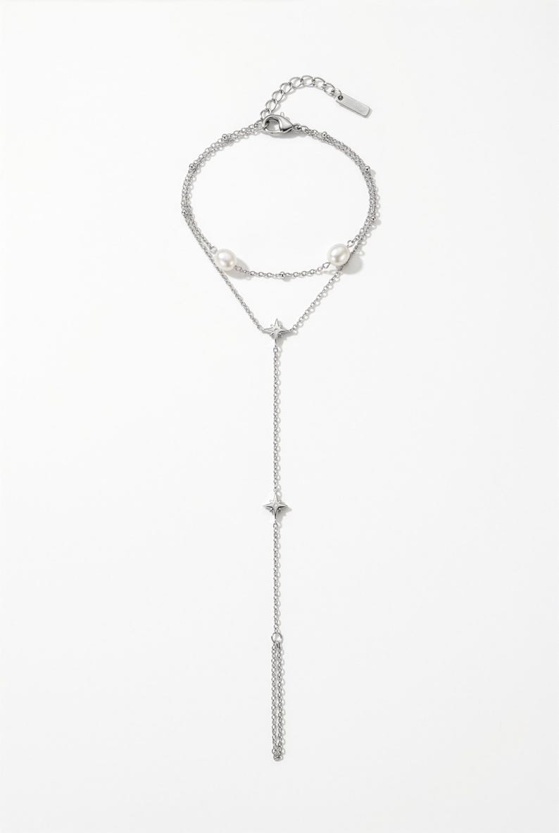 May include: A silver-toned multi-strand bracelet featuring two white pearls and two small star-shaped charms. The jewellery piece includes a delicate chain design with a lobster claw clasp, an adjustable extender chain, and a small rectangular metal tag.