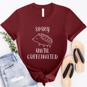 May include: A maroon t-shirt with a white graphic of a hedgehog and the text "Spiky until caffeinated".