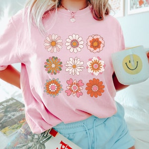 May include: A pink t-shirt with a graphic of nine colorful retro flowers. The flowers are arranged in a grid pattern.