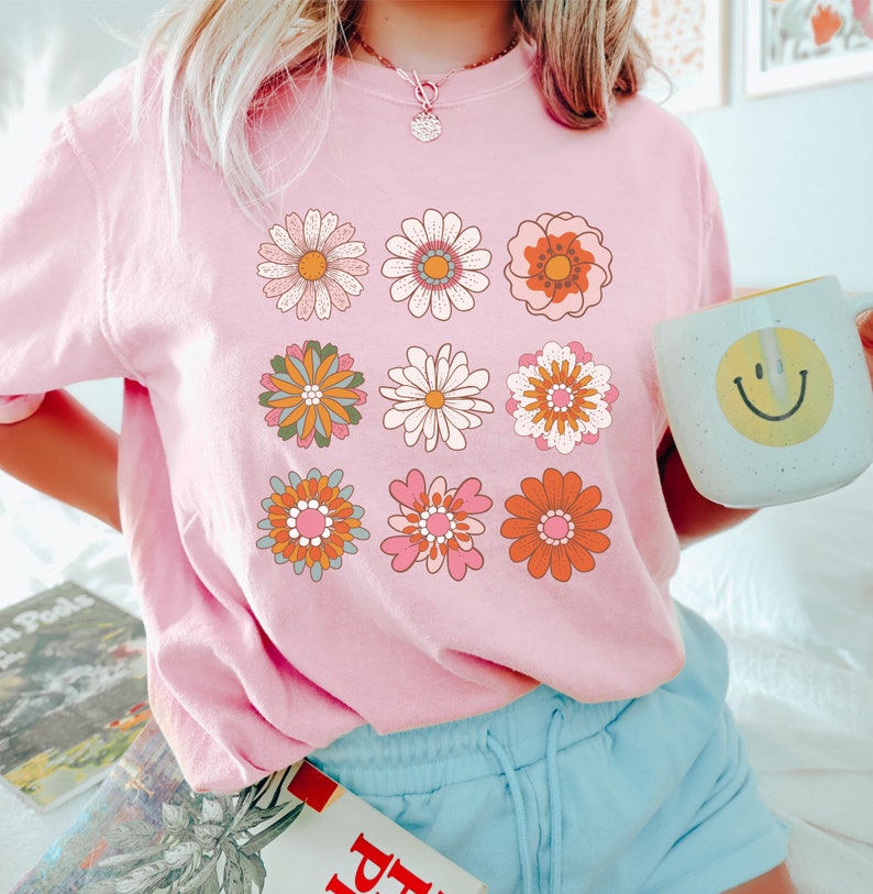 May include: A pink t-shirt with a graphic of nine colorful retro flowers. The flowers are arranged in a grid pattern.