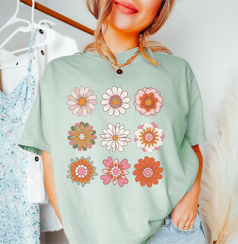 May include: A light green t-shirt with a retro floral design. The design features nine different flowers in shades of pink, orange, yellow, and green.