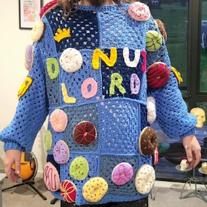 Op de afbeelding: Een blauwe gehaakte trui met een patchwork ontwerp, versierd met talrijke gehaakte donuts in verschillende kleuren. De woorden "DONUT LORD" zijn in kleurrijke letters op de voorkant gespeld. De trui heeft lange mouwen.