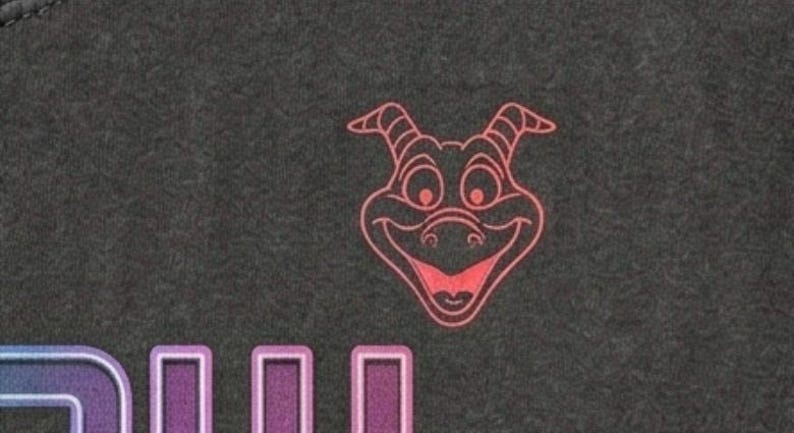 Puede incluir: Una tela gris oscuro que presenta una ilustraci&oacute;n de l&iacute;neas color coral de una cabeza de drag&oacute;n con cuernos y una amplia sonrisa, situada sobre un gr&aacute;fico parcial con letras en degradado morado y azul que tienen un borde blanco.