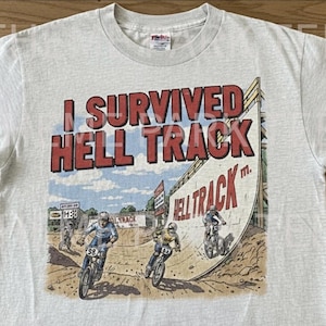 May include: A light gray t-shirt with the text "I Survived Hell Track" in red letters. The graphic depicts BMX riders on a dirt track with the words "Hell Track" on the track.
