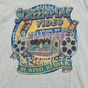 May include: A light gray t-shirt featuring a colorful graphic for "Screenplay Video" in a retro style. The design includes film reels, a storefront, and the text "Be Kind. Rewind."