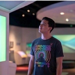 Puede incluir: Una camiseta de manga corta gris oscuro con un estampado gr&aacute;fico de una pir&aacute;mide de colores del arco&iacute;ris con l&iacute;neas circulares conc&eacute;ntricas y texto, usada en una habitaci&oacute;n iluminada con grandes paneles que muestran degradados.