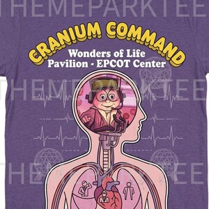 May include: Purple t-shirt featuring the Cranium Command attraction from the Wonders of Life Pavilion at EPCOT Center. The design includes a cartoon character inside a head, with text that reads "Cranium Command" and "Piloted by General Buzzy".