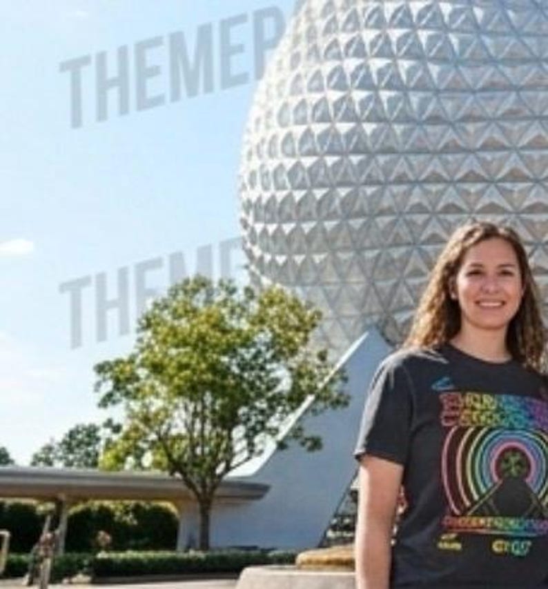 Puede incluir: Una camiseta gris oscuro de manga corta con un estampado gr&aacute;fico colorido de arcos de arco&iacute;ris conc&eacute;ntricos y una silueta de monta&ntilde;a con el texto "THUNDER MOUNTAIN" y "DISNEY" visible en el frente, frente a un gran domo geod&eacute;sico.