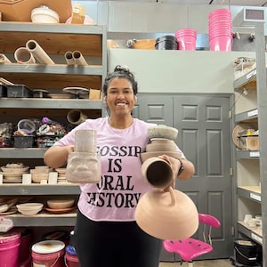 May include: A person holding several pieces of pottery, including a vase and bowls, in a workshop setting. The person is wearing a pink t-shirt with the text "Gossip Is Oral History". Shelves in the background are filled with pottery and supplies.