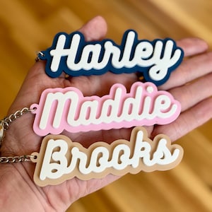 May include: Three personalised keychains with the names "Harley", "Maddie", and "Brooks" in white lettering on a blue, pink, and beige background.