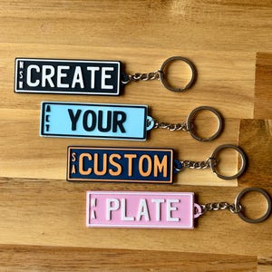 May include: Four rectangular keychains with metal rings. The top is black with white text reading NSW CREATE. The second is light blue with black text reading ACT YOUR. The third is dark blue with orange text reading SA CUSTOM. The bottom is pink with white text reading WA PLATE.