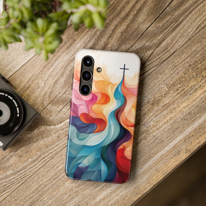 May include: A phone case with a colorful abstract design featuring a cross at the top. The design is made up of swirling lines and shapes in shades of blue, red, yellow, and orange.