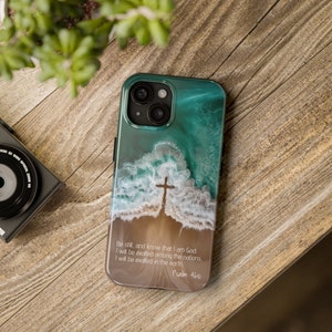 May include: A teal and brown phone case with a white cross on a beach scene. The case has the text "Be still, and know that I am God: I will be exalted among the nations, I will be exalted in the earth Psalm 46:10".