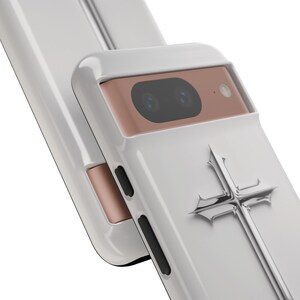 May include: White phone case with a silver cross design and a rose gold camera housing.