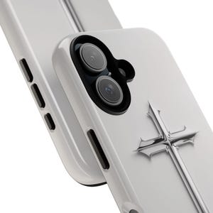 May include: White phone case with a silver cross design. The case has a camera lens cutout and a raised edge around the screen.