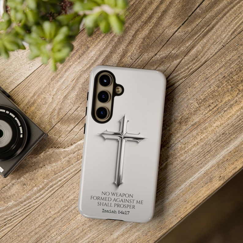 May include: A white phone case with a silver cross and the text "NO WEAPON FORMED AGAINST ME SHALL PROSPER Isaiah 54:17".