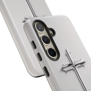 May include: A white phone case with a silver cross design. The case has a black camera bump with three lenses.