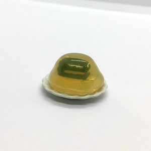 May include: A miniature clear yellow jelly mold with a green layer inside, sitting on a white plate with a scalloped edge.