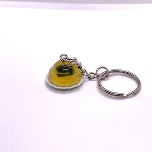 May include: A novelty keychain featuring a miniature yellow dessert encased in clear resin, resting on a white plate. The keychain has a silver-colored chain and a split ring for attaching keys. The dessert appears to have a dark green filling.
