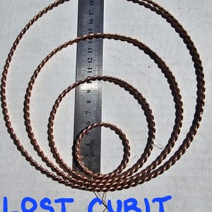 Op de afbeelding: Vier koperkleurige tensorringen van verschillende maten, gerangschikt op een wit oppervlak. Een liniaal is naast de ringen geplaatst voor de schaal. De tekst "LOST CUBIT TENSOR RINGS" staat in het blauw onderaan de afbeelding.