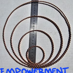 Puede incluir: Cuatro anillos de alambre trenzado de color cobre de varios tamaños, dispuestos concéntricamente. Una regla con marcas de centímetros se coloca junto a los anillos. El texto "EMPOWERMENT CUBIT TENSORS" está escrito en azul en la parte inferior.
