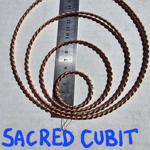 Pode incluir: Quatro anéis tensores Sacred Cubit de cor cobre de vários tamanhos, com uma régua para escala. Os anéis têm um design de corda torcida. O texto "SACRED CUBIT TENSOR RINGS" está escrito em azul na parte inferior da imagem.