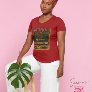 May include: A red t-shirt with a graphic featuring the text "Juneteenth" and "Hey Not Like Us". The shirt has a crew neck and short sleeves. The image also shows a woman wearing a headscarf and white pants.
