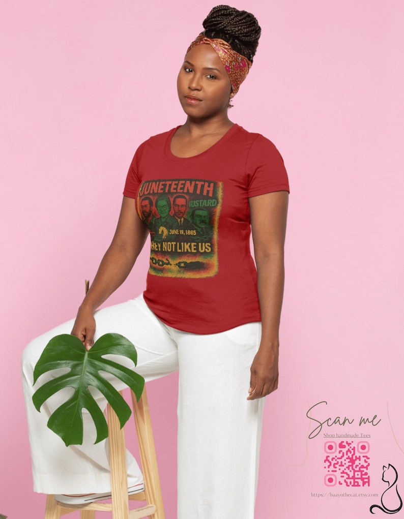 May include: A red t-shirt with a graphic featuring the text "Juneteenth" and "Hey Not Like Us". The shirt has a crew neck and short sleeves. The image also shows a woman wearing a headscarf and white pants.