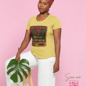 May include: A woman wearing a yellow t-shirt with a graphic that reads "Juneteenth" and "Hey Not Like Us." The shirt features a vintage-style design with portraits and text. She is wearing white pants and a patterned headscarf.