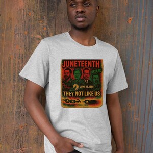 May include: A gray t-shirt featuring a graphic with the text "Juneteenth" and "They Not Like Us." The graphic includes images of four men and the date June 19, 1865. The shirt is worn by a person.