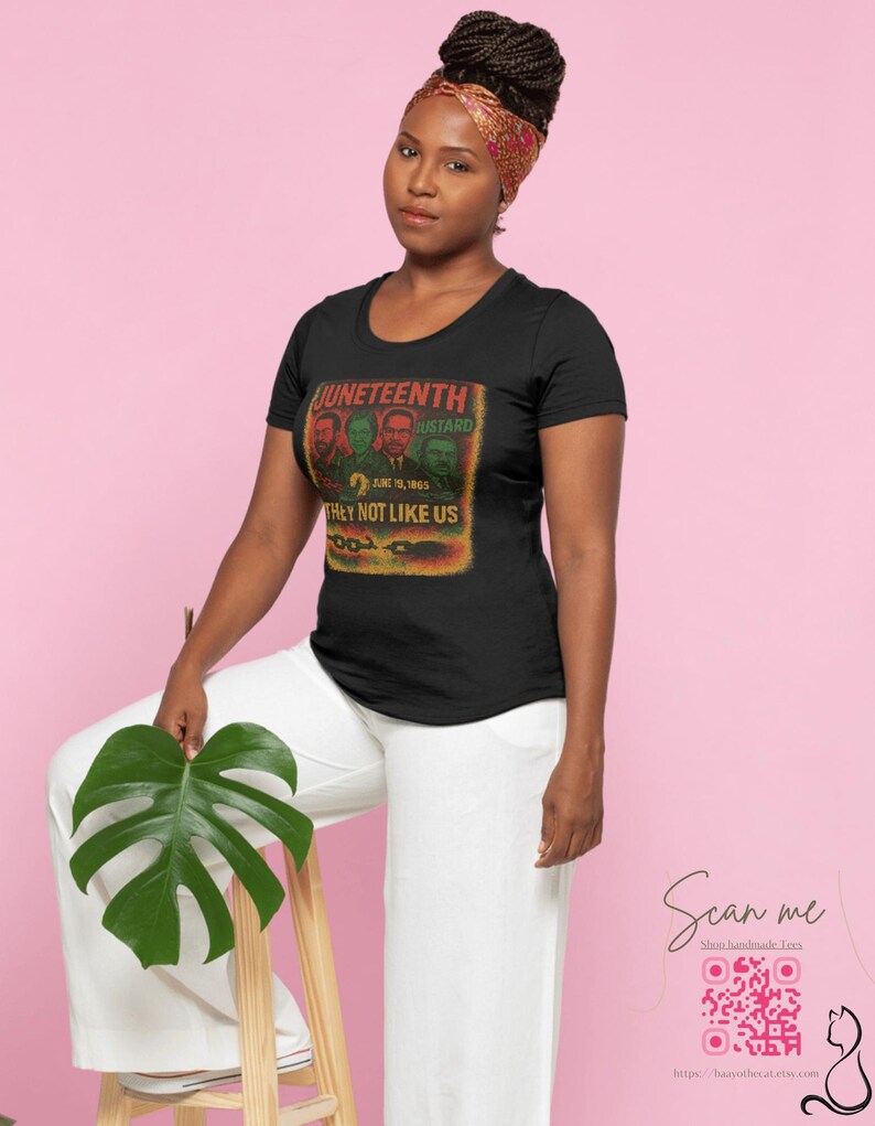 May include: Black t-shirt with a graphic featuring the text "Juneteenth" and the phrase "They Not Like Us." The design includes portraits and the date June 19, 1865. The woman is wearing a patterned headscarf and white pants.