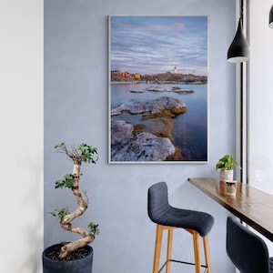 May include: A framed photograph of a seascape with a lighthouse and red houses on a rocky shore. The photo is hanging on a light blue wall above a wooden bar table with two blue bar stools.