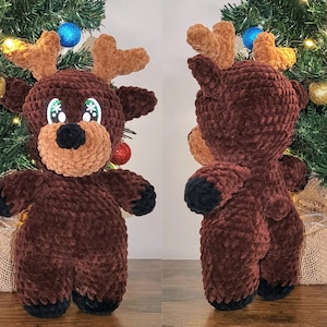 May include: A handmade, plush reindeer toy in shades of brown, with a light brown muzzle and antlers, and black hooves. The reindeer has large, expressive eyes with snowflake accents. It is displayed in front of a small Christmas tree.