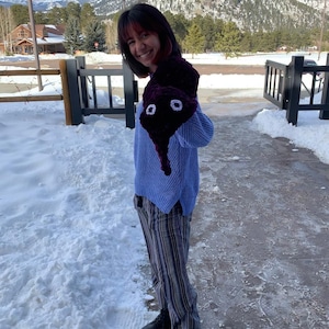 May include: A woman wearing a blue sweater and striped pants is holding a purple crocheted worm with white eyes. The worm is attached to her sweater sleeve. She is standing in front of a snowy background.