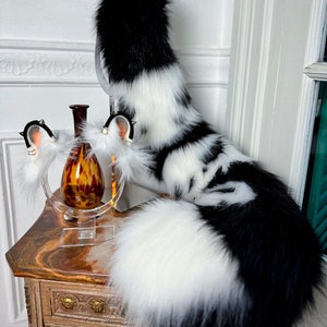 May include: A black and white faux fur tail and matching ear headbands. The tail is striped and fluffy, and the ear headbands have black accents. The items are displayed on a wooden table with a decorative vase.