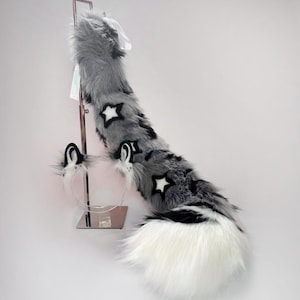 以下が含まれることがあります： 黒い星のアクセントと白い先端が付いた、グレーと白のフェイクファーの尻尾。猫耳付きのお揃いのヘッドバンドも含まれています。 尻尾は金属製のスタンドに展示されています。
