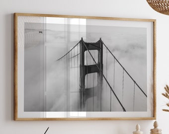 May include: A black and white photograph of the Golden Gate Bridge in San Francisco, California. The bridge is partially obscured by fog. The photograph is framed and hanging on a white wall above a wooden cabinet with woven rattan doors.