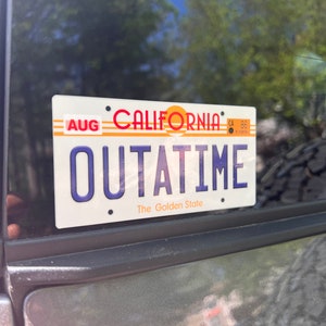 May include: California license plate with the text "OUTATIME" in blue letters. The plate has a white background with red and orange accents, and the words "The Golden State" are at the bottom. The month "AUG" is visible on the left.