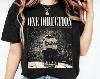May include: Black t-shirt with a white graphic of the band One Direction. The graphic shows the band members hugging on stage in front of a crowd.