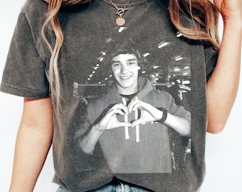 May include: A dark gray t-shirt with a black and white image of a person making a heart shape with their hands. The person is wearing a gray hooded sweatshirt. The t-shirt has a distressed look.