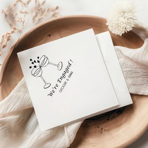 May include: A white card with black ink announcing "We're Engaged!" with a drawing of two champagne glasses clinking and hearts floating above them. The names "George & Diana" are printed below the announcement.