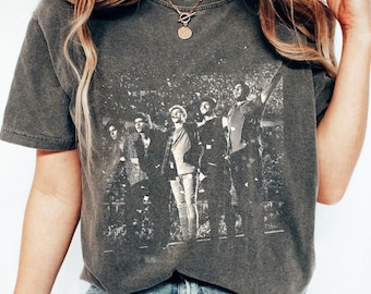 May include: A dark gray t-shirt with a black and white image of a band performing on stage. The band members are wearing casual clothing and are holding microphones. The image is surrounded by a crowd of people.