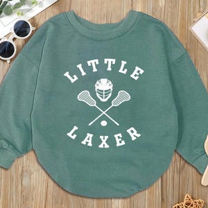May include: A teal green baby bodysuit with the words "LITTLE LAXER" in white block letters. A lacrosse helmet and crossed lacrosse sticks are printed in white below the text. The bodysuit has long sleeves and a round neckline.