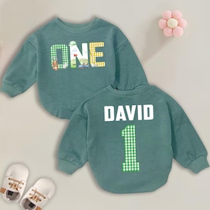 May include: A teal-colored baby sweatshirt with the word "ONE" in a mix of green and yellow checkered patterns and golf-themed illustrations. The back of the sweatshirt has the name "DAVID" and the number "1" in a green checkered pattern.