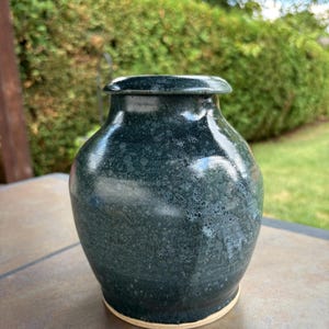 May include: A handcrafted, dark teal ceramic vase with a speckled texture and a glossy finish. The vase has a rounded body, a narrow neck, and a slightly flared rim. The base is a light beige colour.