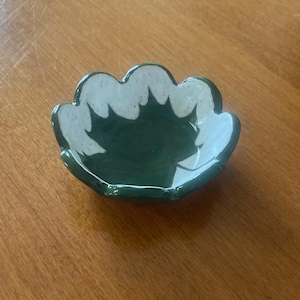 May include: A small, green ceramic dish with a white, scalloped rim. The dish has a white leaf design in the centre.