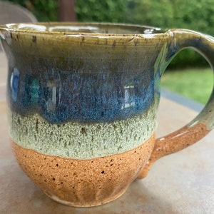 May include: A handmade ceramic mug with a brown, green and blue glaze. The mug has a speckled brown base and a rounded shape.