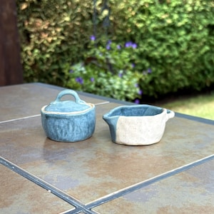 May include: Two small, handmade ceramic containers. One is a lidded pot in a mottled blue glaze. The other is a small, open bowl with a white and blue colour block design. Both are on a tiled surface.